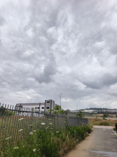 The image shows an outdoor scene in Jezreel Valley Regional Council, Israel, characterized by a grey, overcast sky dominating the upper portion. Below, a grey metal fence runs horizontally across the mid-ground, separating the viewer from a partially constructed or unfinished building with a white and grey facade. Tall weeds and green foliage grow densely along the fence line on the viewer's side. A narrow asphalt path or road runs alongside the vegetation, angling slightly toward the right. In the distance beyond the fence, a grassy area extends toward a low-lying hill with sparse vegetation and buildings. Two street lights are visible, extending vertically in the mid-ground.