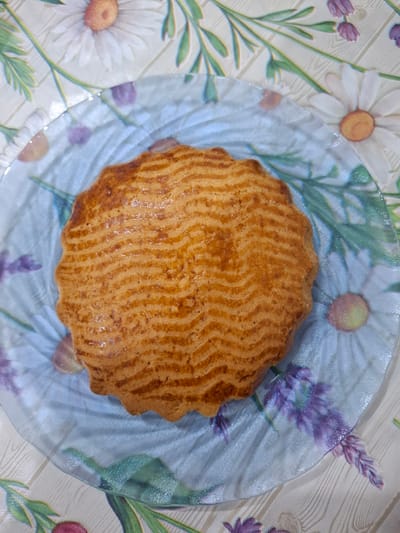 A circular, golden-brown cake with a wavy patterned crust sits on a clear glass plate. The plate is decorated with a floral pattern of white daisies and purple lavender sprigs against a light, wood-grained background. The cake itself has a slightly ruffled edge. The image is taken from a top-down perspective. The setting appears to be indoors, possibly on a table, given the tablecloth-like background pattern. No people are visible, and no text is present in the image. The lighting suggests it could be daytime.