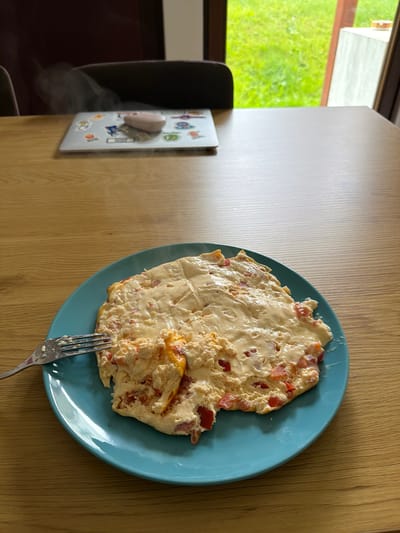 The image shows a close-up view of an omelet with cheese and diced red peppers, served on a blue plate, situated on a wooden table. A fork is partially inserted into the omelet. In the background, a laptop with stickers and a small pink object is visible, with steam rising from it, suggesting it might be hot. Beyond the table, a window reveals a lush green outdoor environment with trees and grass, hinting at a natural setting. The scene appears to be indoors, likely in a home or dining area, with natural light illuminating the table and the food. The overall impression is that of a casual meal being enjoyed.