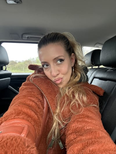 A woman with blonde hair is sitting in the back seat of a car. She is wearing a rust-colored, textured jacket with leather accents and a silver zipper. She has an earring in her left ear. The car's interior is mostly black, and the window shows a view of green vegetation and a gray sky, suggesting an overcast day. The location may be inferred to be South Ockendon, United Kingdom.