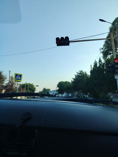 This media file captures a view from inside a vehicle, looking out through the windshield at a street scene during what appears to be late afternoon or early evening due to the warm, soft light. The sky is a clear, pale blue, suggesting fair weather.

Dominating the view are traffic signals and streetlights. A horizontal traffic light with all three lights illuminated red is visible, suspended over the road. To the right, a vertical traffic signal also shows a red light. A pedestrian crossing sign, a blue square with a white pictogram of a person walking on a striped path, is prominent on the left side of the road. Trees line the street, with lush green foliage on the right and sparser, drier vegetation on the left. In the distance, buildings are visible, some appearing to be residential or commercial structures.

Several vehicles are present on the road, including cars and a white truck. The activity depicted is simply traffic moving along a street, with traffic signals indicating a stop. The perspective from inside the car, with part of the dashboard visible, suggests the media was captured by someone driving or a passenger.

A piece of paper with some visible text is attached to a utility pole on the right. The text is in a non-Latin script, likely Cyrillic, and includes numbers such as "33, 927-41". A sign that says "AKBO" is also partially visible. The location context provided is Kurbanov, Uzbekistan. No specific emotions are conveyed, but the scene is peaceful and ordinary.