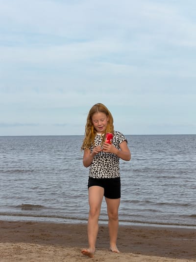 The image depicts a young girl standing on a sandy beach with the ocean in the background. She has long blonde hair and is wearing a black and white leopard print top and black shorts. The girl is holding a red can in her hands and appears to be looking down at it with her eyes closed, suggesting she might be drinking from it or examining it. The water is calm, with gentle ripples on the surface, and the sky is a pale blue with some wispy clouds. The lighting suggests it is daytime, possibly late afternoon or early evening due to the soft light. The presence of sand and the ocean indicates a coastal or lakeside location. The overall mood of the image is relaxed and peaceful. There is no visible text in the image.