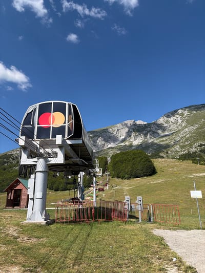 The image depicts a sunny day scene at the Motički gaj ski resort in Montenegro, with a clear blue sky and white clouds. In the foreground, a ski lift station with a cabin featuring the Mastercard logo is prominently visible. The cabin is supported by a large grey pillar. To the left of the station, a small wooden building with a green roof is situated. Red metal fences are arranged around the ski lift area. A path made of gravel leads into the frame from the right. The background features a lush green mountainside with scattered trees and a rocky mountain peak. Several ski lift chairs are visible ascending the slope. The overall environment suggests a warm, late spring or summer day at a mountain resort.