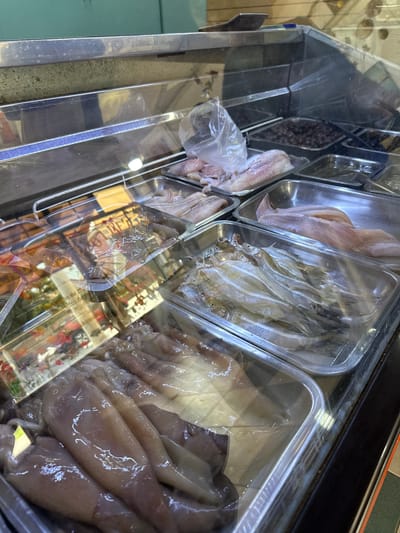 A refrigerated display case containing various raw seafood items in plastic containers is shown. From left to right, the containers hold small, dark items (possibly olives or anchovies), fish fillets, small whole fish, and squid. The display case is located in Villa General Belgrano, Argentina. The environment is a marketplace or food retail establishment.