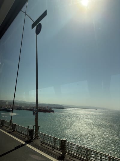 The image displays a sun-drenched, expansive view over a large body of water, likely a bay or sea, from an elevated perspective, possibly a bridge or coastal highway. The bright sun, located in the upper left, casts a strong glare and lens flare, illuminating a clear, light blue sky that dominates the upper half of the frame.
To the right, a road with a robust metal railing runs along the edge of the scene, extending into the distance. The water below glitters with countless reflections of sunlight, indicating calm conditions. Along the distant horizon, rolling hills or mountains are visible, while numerous ships, including large cargo vessels and smaller boats, dot the water. A port or industrial area with cranes and other structures can be discerned on the far shore, suggesting significant maritime activity.
A tall, dark utility pole stands prominently in the foreground, positioned slightly left of center. Attached to this pole is a blue rectangular sign that displays a white phone icon and the text "ACİL YARDIM" above "SOS." This visible text is Turkish for "Emergency Help." Reflections, possibly from a vehicle's window, are subtly visible across the image, particularly in the sky and water areas.
The scene depicts a clear, bright day, likely late morning or early afternoon, given the sun's position. The presence of a bridge-like structure, a busy bay with industrial elements, and the Turkish text on the sign strongly align with the provided context of Dilovası, Turkey.