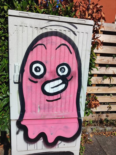 The image shows a pink cartoon ghost with large white eyes and a wide, open mouth, spray-painted on the side of a utility box. The utility box is light gray with vertical textured lines. To the left of the utility box are dark green, glossy leaves of a hedge. To the right is a wooden fence made of horizontal planks, with some dry brown leaves and green plants growing around it. The ground in front of the fence is a paved surface with fallen leaves and some litter. The graffiti covers a significant portion of the utility box, which is located outdoors, likely in a public area. The lighting suggests it is daytime with bright sunlight. There are no people visible in the image. Based on the visible text "Sektfom" which is likely a misspelling of "Sektkellerei" (sparkling wine cellar) and "Verbotene" (forbidden), this suggests the utility box might be related to a business or a former business. Offenburg, Germany is known for its wine production, and it is plausible to find utility boxes of this nature in such a context.
