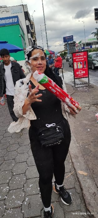 A person in black leggings and a black top lies on a gray stone-paved ground. The person has white foam on their face and hair, holding a red and green foam spray can. A black purse hangs on their side. In the background, there are buildings, street signs, and other people, including one person wearing a white shirt. The picture also shows a date stamp in the upper right corner.
