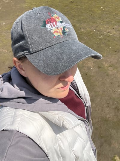 A person, likely female, is shown from the chest up, wearing a gray baseball cap with a floral design and white text that reads "All Saints 2022". They are also wearing a maroon shirt under a gray hoodie, which is then covered by a white puffer vest. The person is looking down and to the right, with their face partially obscured by the cap's brim and shadows. The background is a muddy, unpaved area, possibly a field or shoreline, with some sparse vegetation. The lighting suggests it is daytime. The location is identified as Tullycullion, Ireland.