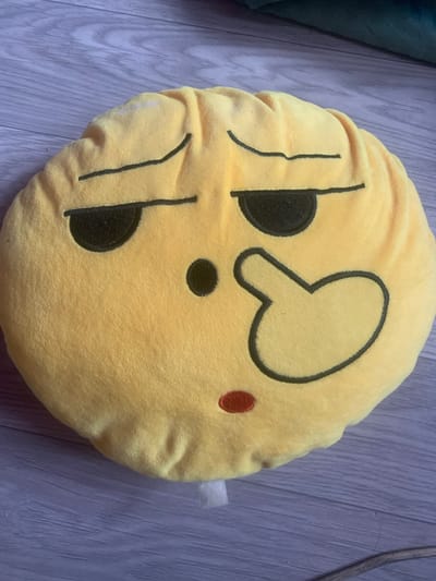 A yellow plush emoji pillow is lying on a wooden floor. The pillow features embroidered facial details: two slanted black eyes, a small black dot for a nose, a hand gesture with a pointer finger extended to the right, and a small red mouth. The background consists of a gray-toned wooden floor with visible planks. There are no people or animals present in the image. The scene appears to be indoors, likely in a residential setting. There is no information about its location in Lebedinovka, Kyrgyzstan.
