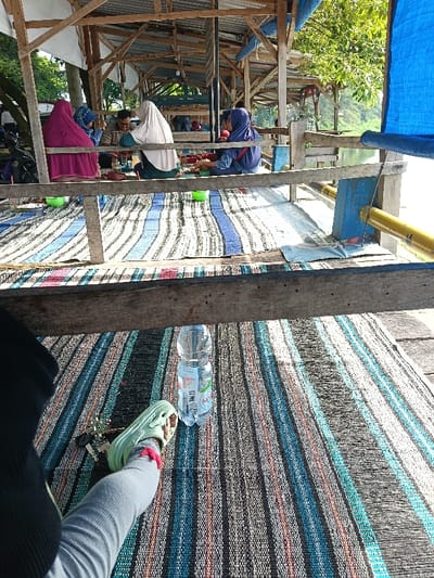 The image depicts an open-air, multi-sectional wooden pavilion structure situated by a body of water in Gembong, Indonesia. The foreground displays the lower left leg and foot of an individual, clad in light gray pants and a light green sandal with a pink ankle strap. A set of keys, including a car key fob, rests on a striped woven mat next to the foot. To the right, a clear plastic bottle of mineral water stands on the same mat.
A horizontal wooden beam spans the mid-frame, separating the foreground from the background activity. Beyond this beam, five individuals are seated on striped woven mats that cover the wooden floor of the pavilion. From left to right: one individual wears a magenta hijab; another wears a blue patterned hijab; a male with glasses in a striped shirt faces right, engaged with an individual in a cream-colored hijab and dark green garment, who is mostly facing away; and a fifth individual wears a blue hijab. Several small green plastic bowls and other containers are distributed on the mats among the seated individuals.
The pavilion is constructed from untreated wooden beams and posts, supporting a roof structure that is partially visible. To the far right, a blue tarpaulin is partially rolled up. Beyond the pavilion, a calm body of water reflects bright sunlight, with the opposite bank lined by green foliage and trees. A portion of a black motorcycle is visible on the far left, behind the seated group. The scene is brightly illuminated by natural daylight.