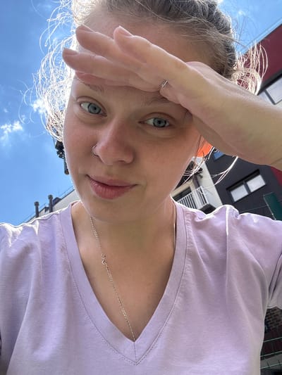 A young Caucasian woman is pictured in an outdoor urban setting, with buildings and a bright blue sky in the background. She is looking directly at the camera with her right hand raised to her forehead. Her blonde hair is pulled back, with wisps of hair illuminated by the sun. She is wearing a light purple V-neck t-shirt and a silver necklace. A nose ring is visible on her left nostril, and a thin silver ring is on her right ring finger. The overall scene suggests a casual, sunny day. The specific location in the city of Vyazniki, Russia, cannot be determined from the visual information provided.