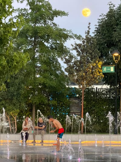 The image depicts an outdoor scene with a prominent full moon in the twilight sky. In the foreground, a young boy wearing red and blue swim trunks stands in a water fountain, splashing water with his hands. To his left, two adults, a woman wearing a headscarf and patterned top, and a man in a dark t-shirt, are seated on a bench. They appear to be observing the boy or the fountain. The fountain area features multiple water jets. Behind the people and the fountain is a hedge and trees. A lamppost with a lit fixture is visible on the right side of the image, along with a green emergency exit sign. A sign with text is also present near the hedge. The overall atmosphere suggests a recreational space in a city. The image was taken in the Gran Plaza 2 in Majadahonda, Spain.