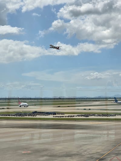 The image depicts an airport scene in Bao'an District, China, under a partly cloudy sky. In the upper portion of the frame, a commercial airplane, predominantly white with red accents, is ascending into the sky. Below, on the tarmac, another similar airplane is visible, appearing to taxi or hold position. Rows of baggage carts are neatly arranged on the ground. In the background, a hazy industrial skyline with cranes and buildings stretches across the horizon. The foreground shows the reflective surface of the airport tarmac.