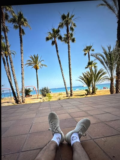 The image shows a low-angle, first-person perspective of a person's legs and feet resting on a tiled surface. The person is wearing light-colored sneakers and white socks. The background features a sunny beach scene with several palm trees, the ocean, and a clear blue sky. There are some beach umbrellas and people visible in the distance. The location is identified as being in the city of Porrentruy, Switzerland, although this scene is inconsistent with the typical geography and climate of Porrentruy, which is an inland municipality.