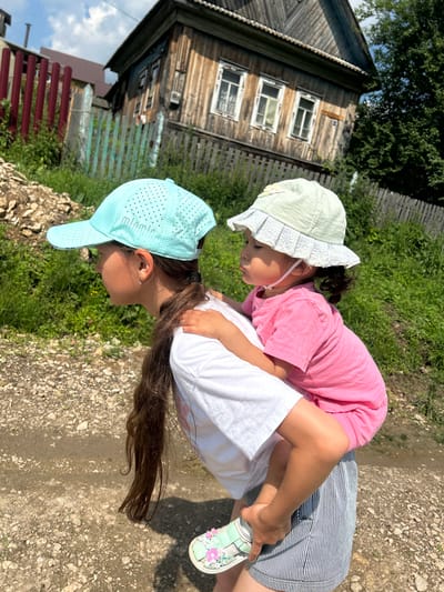 In Новомуллакаево, Russia, on a sunny day, a young girl with long dark hair, wearing a white t-shirt and striped shorts, carries a toddler on her back. The toddler, wearing a pink t-shirt and a light green hat with lace trim, looks to the side. The girl wears a light blue perforated baseball cap. They are walking on a dirt path next to a grassy area, with a weathered wooden house and a rustic fence in the background.
