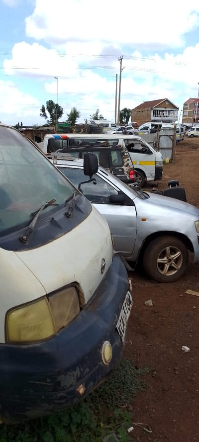 The scene depicts an outdoor vehicle parking area situated in Kuku ward, Kenya, under a cloudy sky. In the immediate foreground, the front section of a weathered light-colored Nissan van is visible, featuring a blue bumper with the partially visible license plate "KCA 318M" and yellowed headlights. Directly behind this van is a silver sedan or small SUV with multi-spoke alloy wheels, exhibiting a dent on its rear passenger side panel; a black object, possibly a headrest, rests within its rear compartment. Further into the mid-ground, multiple white vans are parked, several displaying a distinctive yellow horizontal stripe indicative of public transport vehicles. Some of these vans appear to be in various states of disrepair. The ground consists of reddish-brown dirt with scattered patches of green foliage and minor debris. In the background, several structures, including multi-story buildings, some appearing incomplete with exposed brickwork and corrugated iron roofs, are visible. A prominent wooden utility pole with multiple power lines and a street light pole rise against the sky. Two small, dark, indistinct human figures are present on a low structure in the far left background. No discernible actions or interactions are clearly visible beyond the static placement of vehicles and structures.