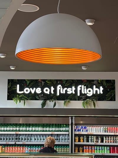 The image shows a brightly lit interior space. A half-dome shaped wall decoration with a white exterior and orange ribbed interior is mounted on a gray wall. Small, round, white lights are situated around it. To the right, a vertical sign with the words "Love at First Flight" in white block letters is set against a backdrop of green foliage. Adjacent to the sign is a vending machine filled with a variety of beverages and snacks. A person with blonde hair is seen partially in the frame, apparently looking at the contents of the vending machine. The image is taken inside Kruja, Albania.