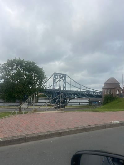 This image captures a view of the Rüstringer Bridge in Wilhelmshaven, Germany, on an overcast day. The bridge, a prominent steel suspension bridge painted a dark blue, spans a waterway. To the left of the bridge, a large, leafy tree stands near a set of metal stairs leading down to a lower level. A brick building with a distinctive brown roof and windows is situated to the right of the bridge, atop a grassy embankment. In the foreground, a paved area with red brickwork and a grey concrete curb borders a road. A section of a car's side mirror is visible in the bottom right corner of the frame. A black car is parked on the road to the left of the bridge, and another black car is further back, on the roadway below the bridge. The sky is heavily clouded.