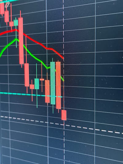 The image is a close-up, low-angle, and slightly blurry photograph of a computer screen displaying a financial trading chart. The chart shows several candlestick patterns in red and green, indicating price movements over time. Red candlesticks generally represent a price decrease, while green candlesticks represent a price increase. Two lines, one red and one green, are also visible, likely representing moving averages or other indicators. The background of the screen is dark, with a grid overlay of thin white horizontal and vertical lines. A dashed white horizontal line is also present. There are no people, animals, or specific landmarks visible, and the image provides no information about the city of Gresham, United States of America. The focus is solely on the abstract visual representation of market data on a digital display.