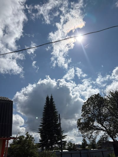 The image shows a view of the sky with clouds, a bright sun, and a power line. In the foreground, there are silhouetted trees, a building with a corrugated roof, and a fence. The location is in the city of Sayansk, Russia. The sky is a vibrant blue with fluffy white clouds, some of which are casting shadows. The sun is high in the sky, and its rays are visible, creating a lens flare effect. A power line runs diagonally across the upper portion of the image. The trees are dense and dark green, and the building appears to be made of dark material. The fence is visible in the lower part of the image. The overall scene depicts an outdoor setting on a sunny day.