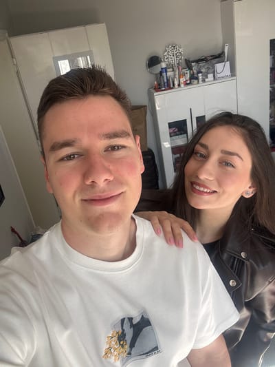 A selfie of a man and woman indoors. The man, with short brown hair, is wearing a white t-shirt with a print on the chest. He is holding the camera and smiling slightly. The woman has long brown hair and is wearing a dark leather jacket. She is positioned behind the man, with her hand on his shoulder, and is also smiling.
The background shows furniture, including a white cabinet with items such as a Pandora box and toiletries. The photo may have been taken indoors in a living room. There is no indication that this image was taken in the city of Bukovlak, Bulgaria.