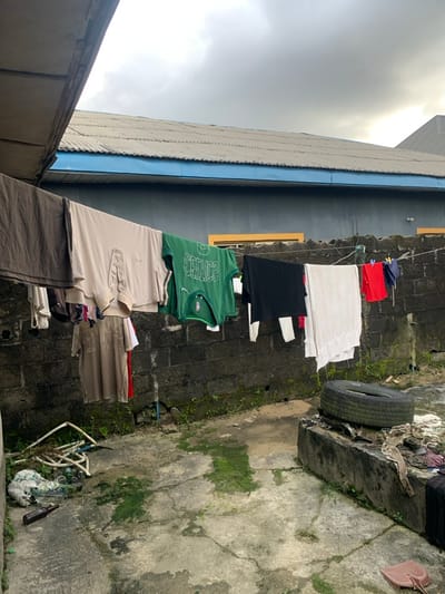 A narrow outdoor space with clothes hanging on a line between two buildings. Various items of clothing, including shirts and towels, are displayed. A tire rests on a concrete ledge along the wall. The ground is concrete, partly covered with moss and miscellaneous debris. The buildings are adjacent and of varying heights, the taller building is dark with a blue trim, while the lower building is lighter in color. The sky is overcast.