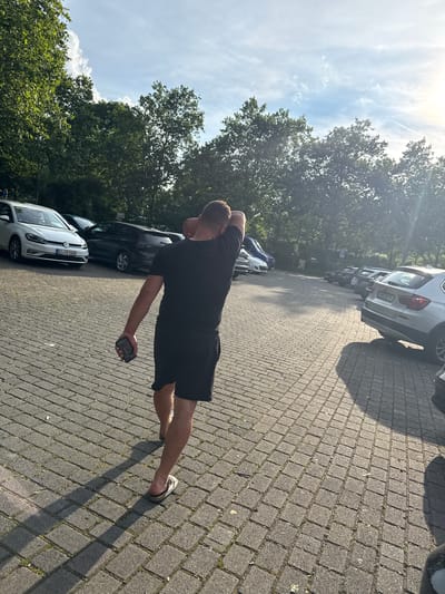 The image captures a sunlit outdoor parking lot in Wetzlar, Germany. In the foreground, a man wearing a black t-shirt and black shorts is viewed from behind, walking away from the camera. He is holding a dark-colored mobile phone in his right hand, and his left arm is raised, with his hand touching the back of his head. He is wearing light-colored flip-flops. The parking lot surface is paved with interlocking grey bricks. Several cars are parked in the lot, including a silver Volkswagen Golf on the left, and a silver BMW SUV on the right. Lush green trees with dense foliage line the background, and the sky is a clear blue with scattered white clouds, indicating a bright, sunny day. The sun is visible in the upper right corner, casting long shadows across the parking lot.