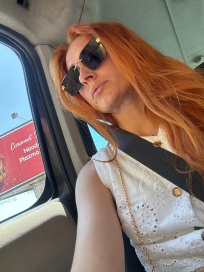 The media file shows a close-up of a person with long, reddish-orange hair, wearing sunglasses and a white, eyelet-patterned top. The person is inside a vehicle, likely a car, as indicated by the car's interior visible in the background and a seatbelt. The person's face is partially obscured by the sunglasses, but their mouth is slightly open, suggesting a relaxed or perhaps candid moment. A gold chain bag strap is visible across the person's chest.
The scene appears to be outdoors, with natural light coming in from a window, casting shadows. A billboard is visible through the car window, with text that reads "Caramel", "Neod", "Plazma". The text is in a mix of red and white, with the word "Caramel" in a flowing script font.
The overall impression is of a casual moment captured during daytime travel. The lighting suggests it might be a sunny day. The image is framed from a high angle, looking down at the person.