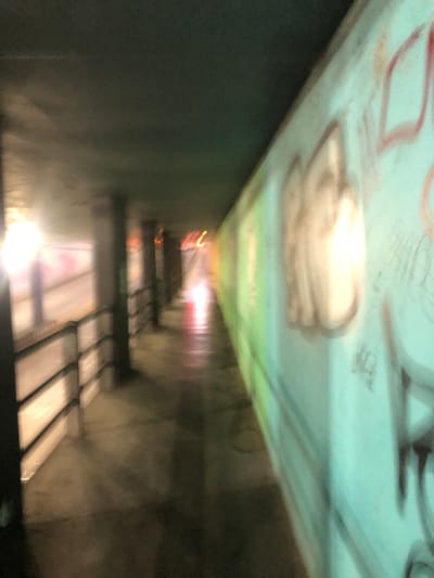 A blurry, low-angle shot depicts a dark, narrow tunnel or passageway. Graffiti covers the ceiling. Light sources exist at varying depths within the tunnel. Stair treads are visible in the lower portion of the frame. There are no people visible. The location is unknown.