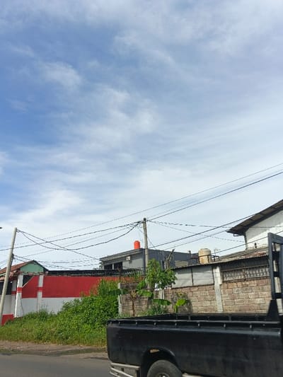 A partly cloudy sky overlooks a street scene. Various buildings and walls are visible, including one painted red and white. An orange water tank sits atop a gray building. A black truck is partially visible in the foreground, along with overgrown foliage. Overhead, electrical wires crisscross the sky.