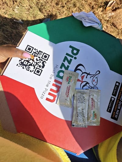 The image captures a close-up, angled view of a "Pizza Inn" pizza box in what appears to be an outdoor setting. The box itself is predominantly white, with red and green sections at the bottom left and top right, respectively, reflecting the colors of the Italian flag. The "Pizza Inn" logo, featuring the brand name in green and a cartoon-like pizza character, is prominently displayed. Below the logo, the slogan "Must be the Pizza" is printed vertically.

A person's hand, with light pink manicured nails, holds the box on the left side. One of the nails has a small, intricate design. A large black QR code is printed on the top left corner of the white section of the box.

Resting on the white and red section of the pizza box are three small, rectangular sachets of "Pizza Inn TOMATO KETCHUP." One sachet clearly shows "Pizza Inn TOMATO KETCHUP" printed on it. Another sachet, positioned centrally, has "MFG.18.10" (manufacturing date) and "EXP. 10. 2025" (expiration date) visible on its bottom edge.

On the far right side of the box, next to the green section, there are icons for the "App Store" and "Google Play," with text encouraging users to "WIN NEW DELIVERY APP!" and mentioning "INN KENYA DELIVERY APP!"

The background of the image consists of dry, earthy ground with hints of withered grass, suggesting a sunny outdoor environment, as indicated by the distinct shadows cast across the pizza box. A crumpled white paper item is visible on the upper green edge of the box. At the bottom left, a bright yellow, textured fabric, possibly a towel or blanket, is partially visible.