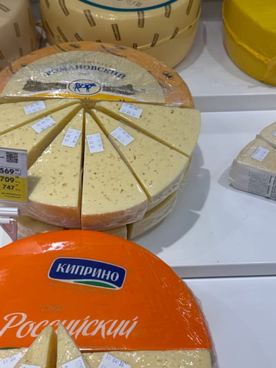 This media file shows a close-up, angled view of several wheels of cheese displayed in a retail setting, likely a grocery store or market. In the foreground, a large, bright orange wheel of cheese is prominent, with some slices cut from it revealing a pale yellow interior. White text in a stylized font is visible on the orange rind, including "КИПРИНО" and "Российский". To the left of the orange cheese wheel, a circular cheese, also sliced into wedges, is wrapped in plastic. This wheel has a label with Cyrillic text that appears to say "РОМАНОВСКИЙ" and features a blue graphic of a cow. Small white labels with dates, such as "25 04 26" and "23 08 26", are affixed to the cheese wedges. In the background, a portion of a large, yellow-wrapped cheese wheel is visible. The setting appears to be indoors with bright, even lighting. The focus is on the cheese products, with no people or specific time of day details evident. The overall impression is a display of dairy products for sale.