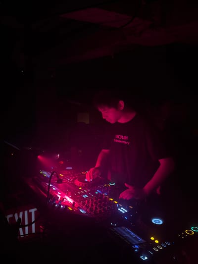 The image depicts a male DJ performing in a dimly lit club or event space located in Changning District, China. The primary illumination is a deep red or magenta light, casting significant shadows and highlighting the subjects and equipment. The central figure is a young adult male with short dark hair, wearing a dark short-sleeved t-shirt with the text "VACUUM 1st Anniversary" printed in pink across the chest. He is positioned in front of a professional DJ console, intently focused on the equipment. His right hand is actively engaged with the circular jog wheel of the rightmost deck, while his left hand rests near the mixer's faders.
The DJ setup consists of multiple decks and a central mixer, featuring numerous illuminated buttons, knobs, and screens in various colors including red, blue, yellow, and white. A small gooseneck lamp extends over the mixer, providing localized light on the controls. In the lower-left foreground, a red 'EXIT' sign with illuminated white letters is partially visible. The background is largely obscured by darkness, though faint, indistinct silhouettes suggest the presence of other individuals deeper within the space. The overall environment is characterized by its low light conditions and the glowing operational lights of the sound equipment.