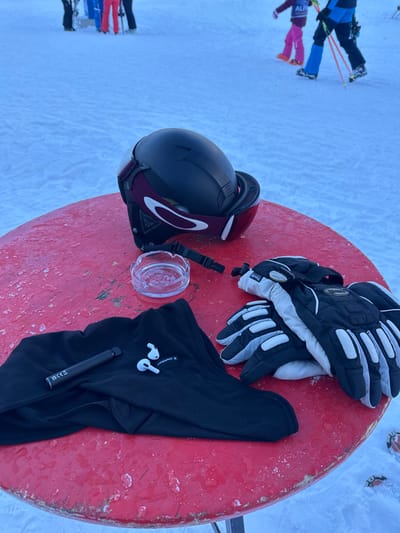The image displays a weathered, round red table situated on a snow-covered ski slope in Bad Hofgastein, Austria. On the table, from left to right, a black balaclava or beanie is laid out, upon which a black vape pen and a pair of white Apple AirPods rest. Adjacent to these items is a clear glass ashtray. To the right, a black ski helmet with an integrated Oakley goggle strap, featuring maroon accents, is placed with its goggle lenses facing upward. Next to the helmet, a pair of black, grey, and white ski gloves, bearing "Thinsulate" and "Kessler" branding, are stacked. In the background, on the snow, several individuals are visible: a person in blue ski attire holding ski poles, a child in pink ski clothing, and other indistinct figures further away. The overall environment is an outdoor, snowy ski area.