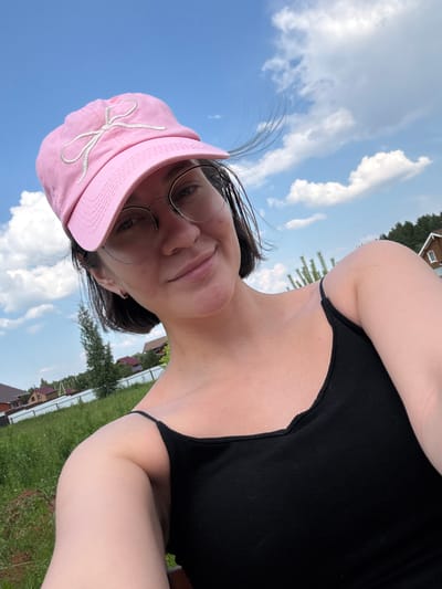A young woman with short, dark hair is taking a selfie outdoors on a sunny day. She is wearing a pink baseball cap with a white embroidered bow and round-rimmed glasses. Her expression is a slight smile. She is wearing a black tank top. The background shows a blue sky with white clouds, green grass, a white fence, and several houses with red roofs, suggesting a suburban or rural setting. Trees and foliage are also visible. The image is likely taken in the city of Dokshitsy, Belarus, not Doksha, Russia.
