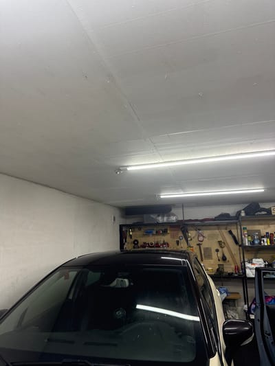 The image shows the interior of a garage or workshop. In the foreground, the roof and windshield of a black car are visible. The car appears to be parked inside the garage. In the background, there are shelves mounted on the wall, filled with various tools, containers, and other items. Two long fluorescent lights are mounted on the ceiling, illuminating the space. The walls are plain and light-colored. The overall impression is one of a functional, albeit somewhat cluttered, workspace. The location is specified as Bolshie Ozeryki, Russia. There are no people or animals visible in the image.