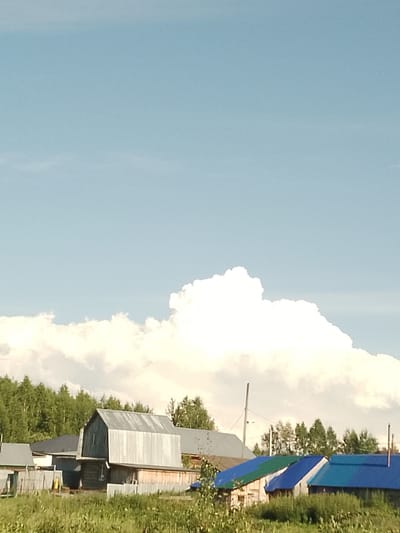 This image depicts a rural scene in Абалак, Russia, under a bright, partly cloudy sky. The foreground is dominated by lush green grass and vegetation, suggesting it's daytime, likely late spring or summer. In the mid-ground, several structures form a village or homestead.
The most prominent building is a rustic, two-story wooden house with a corrugated metal roof that has a distinctive, almost curved, slope. The house has a few windows visible and appears to be constructed from dark wood. To its left, a smaller, single-story building with a grey roof is partially visible, with a wooden fence separating it from the foreground. Further to the right, a row of buildings with brightly colored roofs – blue and green – are lined up, suggesting residential or storage structures. A tall, thin pole, possibly a utility pole or antenna, stands behind these buildings.
The background is a dense line of green trees, characteristic of a forest or wooded area. Above the trees, large, billowy white clouds are scattered across a pale blue sky. The light suggests it's daytime, with the sun high enough to illuminate the scene and create some shadows, although they are not very pronounced. There is no discernible text in the image. The overall impression is one of a peaceful, quiet rural setting on a pleasant day.