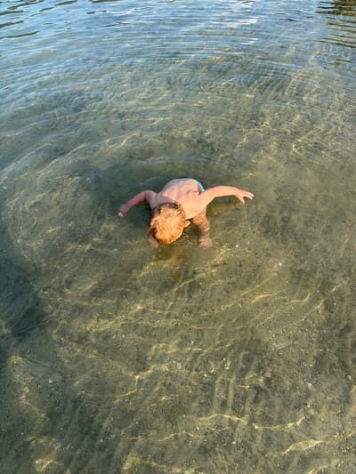 This aerial shot captures a young child with blond hair wading in shallow, clear water. The child, wearing light-colored swim trunks, is facing away from the camera, with their right arm extended forward as if reaching out. The water is so transparent that the sandy, pebble-strewn bottom is clearly visible, illuminated by dappled sunlight that creates shimmering patterns on the surface. The light suggests it's daytime, likely a sunny day given the brightness and reflections. The scene appears to be a calm body of water, possibly a lake or a shallow river, given the gentle ripples. There is no visible text in the image.