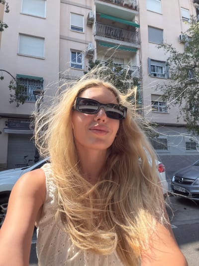 This is a selfie taken outdoors, likely in a European city given the architecture. A young woman with long, blonde hair is the primary subject. She is wearing black, rectangular-shaped sunglasses and a white, eyelet-detailed top. Her hair is windswept, illuminated by bright sunlight. The background shows a building with multiple windows and balconies, characteristic of apartment buildings. To the right, part of a car is visible, with a license plate reading "0257 FVY". There are also some trees and what appears to be a street in the background. The lighting suggests it is daytime, likely sunny. The overall mood is casual and relaxed. A sign with text "El Cafe" is partially visible on a building in the lower left portion of the image.