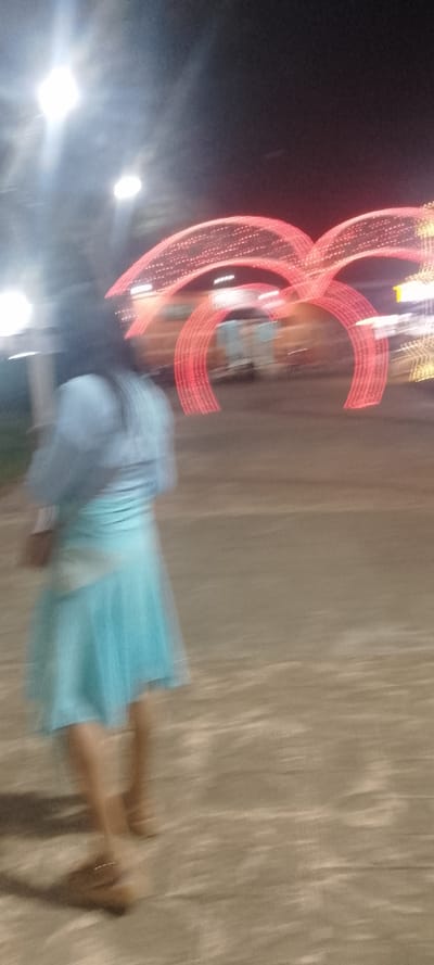 The image depicts a nighttime outdoor scene in La Fría, Venezuela. In the foreground, the blurred figure of an individual with dark, shoulder-length hair is visible from the back, walking away from the camera. The person wears a light blue or teal top and a matching skirt, with brown shoes on their feet. The ground surface is paved and appears light brown or grey. In the midground and background, multiple large, arch-shaped structures illuminated with red lights are prominently displayed. To the right of these arches, additional yellow decorative lights are partially visible, suggesting a festive or holiday display. Overhead, several bright white light sources, consistent with streetlights or floodlights, produce significant lens flare against the dark night sky. The overall image exhibits motion blur, particularly affecting the person and the illuminated structures.