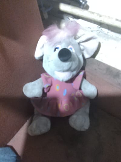The image shows a close-up, slightly blurry view of a stuffed koala toy lying on a reddish-brown surface. The koala is gray with large, prominent ears, a black nose, and blue eyes. It is wearing a pink dress adorned with colorful circular patterns in yellow, blue, and light green. The toy has light pink tufts of fur on its head, resembling hair. Its limbs are visible, including its feet and arms. The background is dark and indistinct, with some lighter vertical elements on the left side, possibly part of the setting. The lighting in the image is dim, and the orientation is somewhat tilted, making it difficult to discern specific details about the environment. The overall impression is of a toy placed on a surface, possibly indoors, with poor lighting conditions. No people, text, or specific time of day or weather cues are discernible.