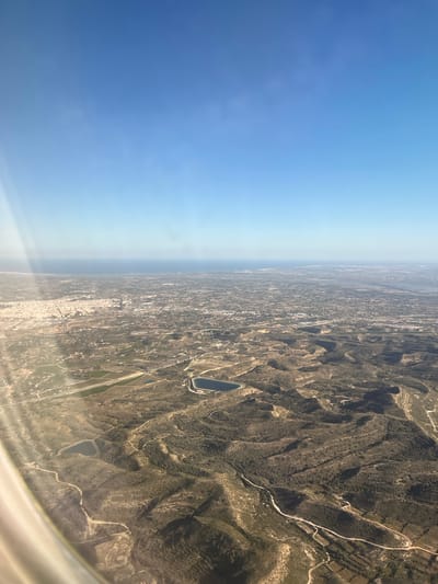 The image captures an aerial view of the landscape near the city of Aspe, Spain, under a clear blue sky. The land is a mix of hilly terrain and flat plains. A small lake is visible amidst the landscape. The area appears to be a mix of natural terrain and some urban development. The photograph appears to be taken from an aircraft.
