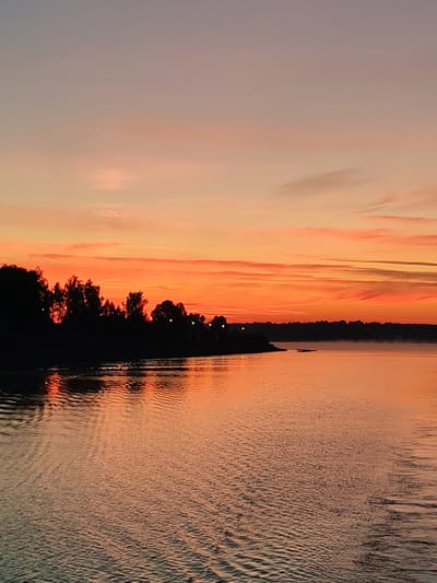 The image depicts a tranquil panoramic view of a large body of water at either sunrise or sunset, specifically in the vicinity of Myshkin, Russia. The upper third of the frame is dominated by the sky, which exhibits a vivid gradient of colors: a muted purplish-blue at the very top transitions to soft peach and then intensifies into brilliant oranges and reds closer to the horizon. Elongated, wispy clouds streak across the sky, catching the light in various shades of orange and red. A dark, silhouetted landmass, densely covered with trees, stretches horizontally across the mid-ground, delineating the horizon. Within the tree line on the left side, faint points of artificial light are visible. On the right side of the distant landmass, a subtle, low-lying mist or haze is present just above the water surface. The lower two-thirds of the image capture the expansive body of water, which vividly mirrors the fiery hues of the sky in shades of orange and red. The water surface is characterized by numerous small ripples and waves, particularly pronounced in the foreground, creating a textured pattern and reflecting fragmented light. No individuals, boats, or other distinct man-made objects are identifiable in the immediate foreground or mid-ground.