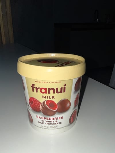 The media file is a close-up, slightly tilted shot of a round, pint-sized container of "Frunui" brand milk chocolate with raspberries. The container is predominantly white with a yellow lid. The design features red raspberries and brown chocolate pieces, along with the brand name "Frunui" in a stylized font. Text on the container indicates it is a "RECIPE FROM PATAGONIA," contains "RASPBERRIES IN WHITE & MILK CHOCOLATE," and has a "Net weight 150g." The image has a somewhat dramatic, low-light setting, with shadows obscuring much of the background, suggesting an indoor environment, possibly a countertop. The focus is sharp on the product itself, highlighting the packaging details. There are no people or animals visible in the image.
