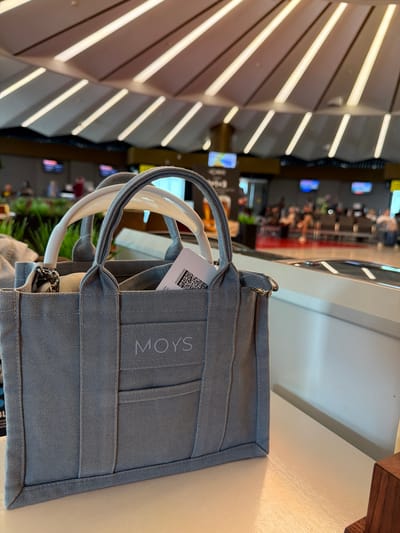 The image shows a close-up of a gray canvas tote bag with the word "MOYS" embossed on it in a lighter gray. The bag has white and gray handles. A tag with a barcode is sticking out of the bag. The bag is placed on a white surface, possibly a table, in an indoor setting with a blurred background. The background appears to be a public space with people sitting in chairs, televisions displaying content, and a ceiling with a distinctive curved design with integrated lighting. The lighting suggests it could be an airport lounge or waiting area, possibly at an airport in or near Перепечино, Russia, given the context provided. There are no visible people interacting with the bag.