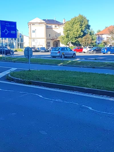 The image depicts a street scene in Nový Jičín, Czechia. A blue directional signpost with white text indicates "57 VSETÍN" and "VALAŠSKÉ MEZIŘÍČÍ," along with a roundabout symbol and the word "Centrum." Parked cars, including a silver hatchback, a red car, a white car, a black car, and a blue car, are visible in a lot or along the street. A portion of a building with light-colored walls and a light brown roof is in the background, as is a large green tree. The foreground shows a road with white lane markings and a grass verge.