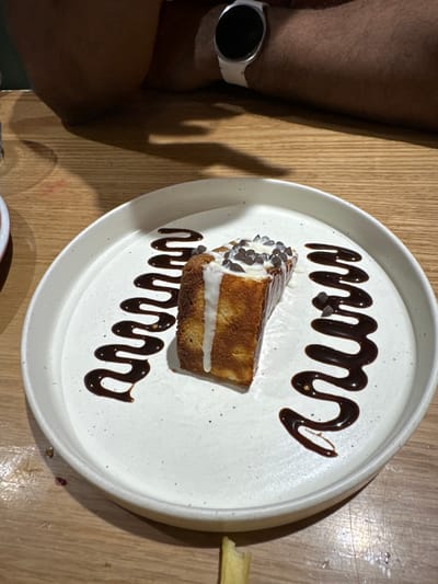 The image displays a dessert presented on a round, off-white ceramic plate with a speckled texture, resting on a light-colored wooden table. The dessert is a rectangular slice of a baked good, possibly cake or bread pudding, topped with a white cream or sauce and numerous small, dark chocolate chips. Two wavy lines of dark chocolate sauce are artfully drizzled on the plate, flanking the dessert. A portion of a person's dark-skinned forearm is visible in the upper background, resting on the table. A white smartwatch with a dark circular screen is worn on the wrist. Shadows are cast across the wooden table surface, suggesting an indoor setting with overhead or directed lighting. A small, indistinct light-colored object, possibly a food remnant like a french fry, is visible on the table surface in the lower foreground. The specific location within Kalyan-Dombivli, India, cannot be determined from the visual information provided.