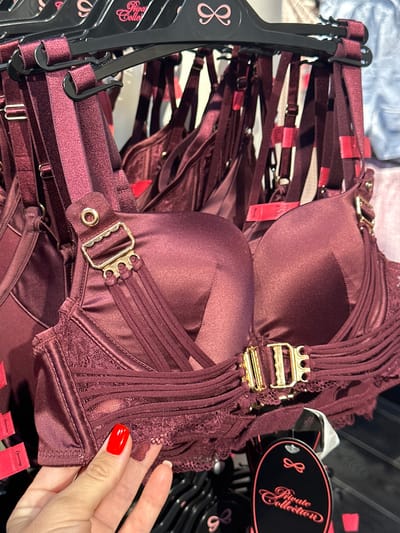 A woman's hand with red nail polish is holding a burgundy satin bra with gold hardware and lace trim, displaying it against a rack of similar bras in a retail environment. The bra features adjustable straps, a balconette cup, and intricate strap details across the bust. The background shows a wall of bras on hangers, some with pink tags featuring a bow design and "Private Collection" text. The lighting suggests an indoor store setting. The image is cropped to focus on the bra and the woman's hand.