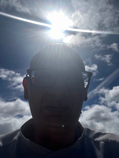 This is a low-angle, close-up, front-facing, selfie-style photograph of a person outdoors under a bright sun and a blue sky with scattered clouds. The sun is directly overhead and is the brightest element in the image, creating lens flare and washing out much of the detail in the upper part of the frame.

The subject, viewed from the chin up, is wearing glasses with dark frames. Their head appears bald. Their skin tone is not clearly discernible due to the strong backlighting. A portion of their ear is visible on the left side of the frame. They are wearing a gray collared shirt or jacket.

The background consists of a vibrant blue sky with fluffy white clouds. There is a faint contrail visible in the upper right quadrant of the sky.

The image does not contain any specific landmarks or details that would allow for the precise identification of a location within Tewkesbury, United Kingdom. It appears to be a generic outdoor scene on a sunny day.