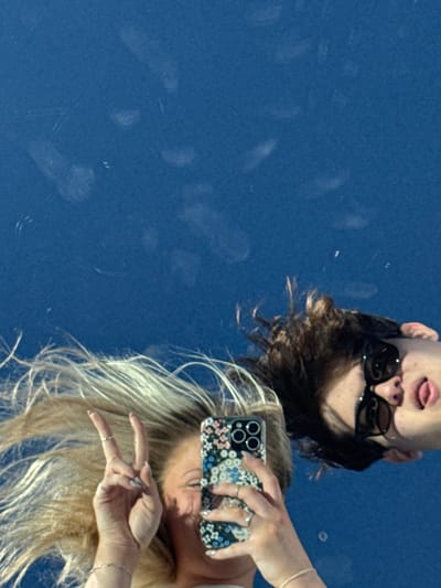 Two people take a selfie, reflected in a shiny blue surface. One person wears sunglasses, sticks out their tongue, and has short brown hair. The other person holds a floral-patterned phone and flashes a peace sign. They have long blonde hair. The surface has smudges and marks, adding texture to the image. The perspective is unusual, as the reflection is inverted, giving a sense of playfulness.